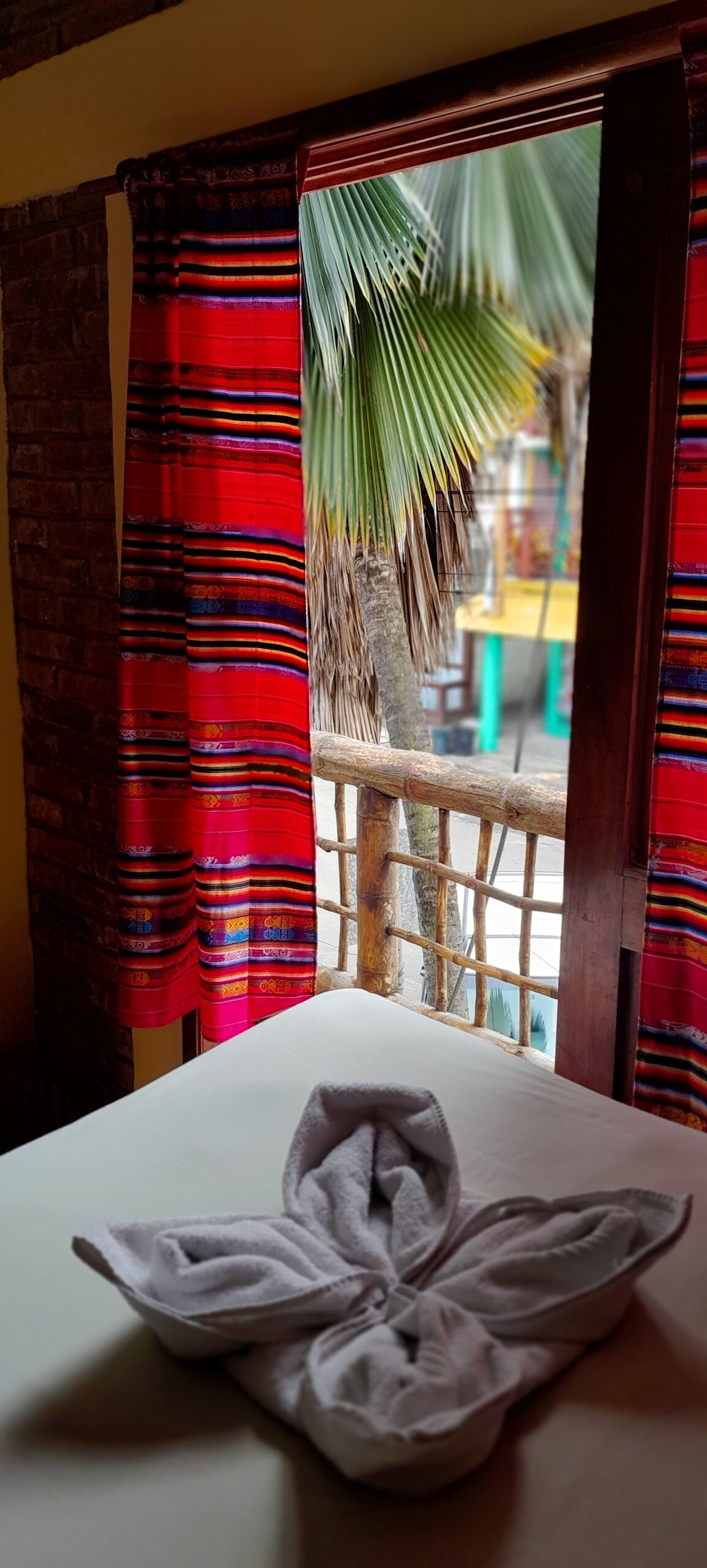 A window adorned with vibrant, multicolored curtains provides a view of palm trees and an outdoor area. A neatly arranged towel in the shape of a flower is placed on the bed, complementing the warm and inviting atmosphere of the room.