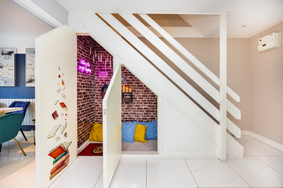 A cozy nook is tucked beneath the staircase, featuring a brick wall and colorful cushions. The entrance is marked by an artistic door with book illustrations, inviting relaxation and playful moments in this charming space.