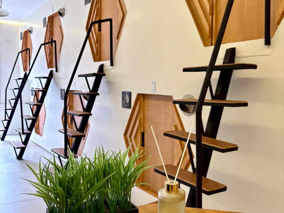 A modern interior features hexagonal pods aligned along a wall, each accessed by sleek black staircases. The scene includes a wooden table with a small plant and a decorative diffuser in the foreground, creating an organized and minimalist atmosphere.