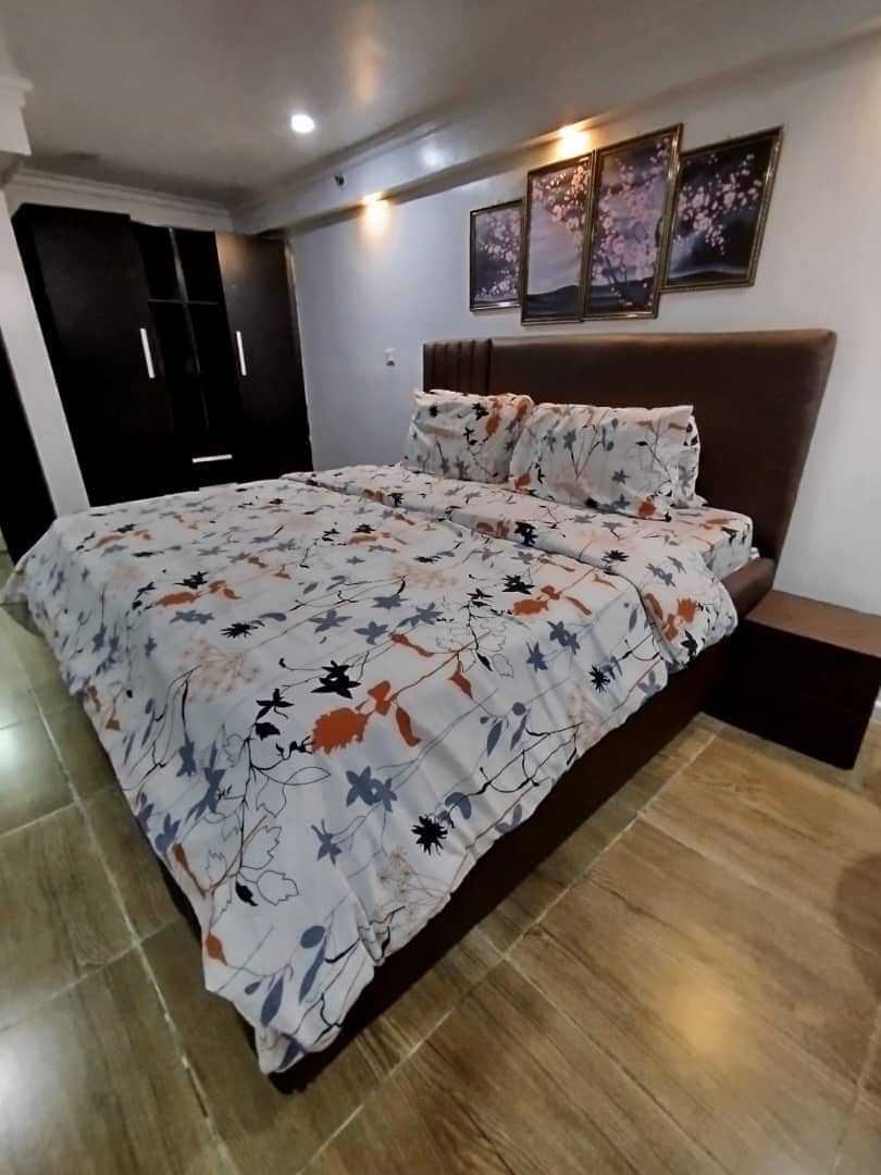 The bedroom features a spacious king size bed adorned with a floral-patterned duvet. Dark wood cabinetry is positioned against the wall, and three framed artworks hang above the bed. Soft lighting enhances the calming ambiance of the room.