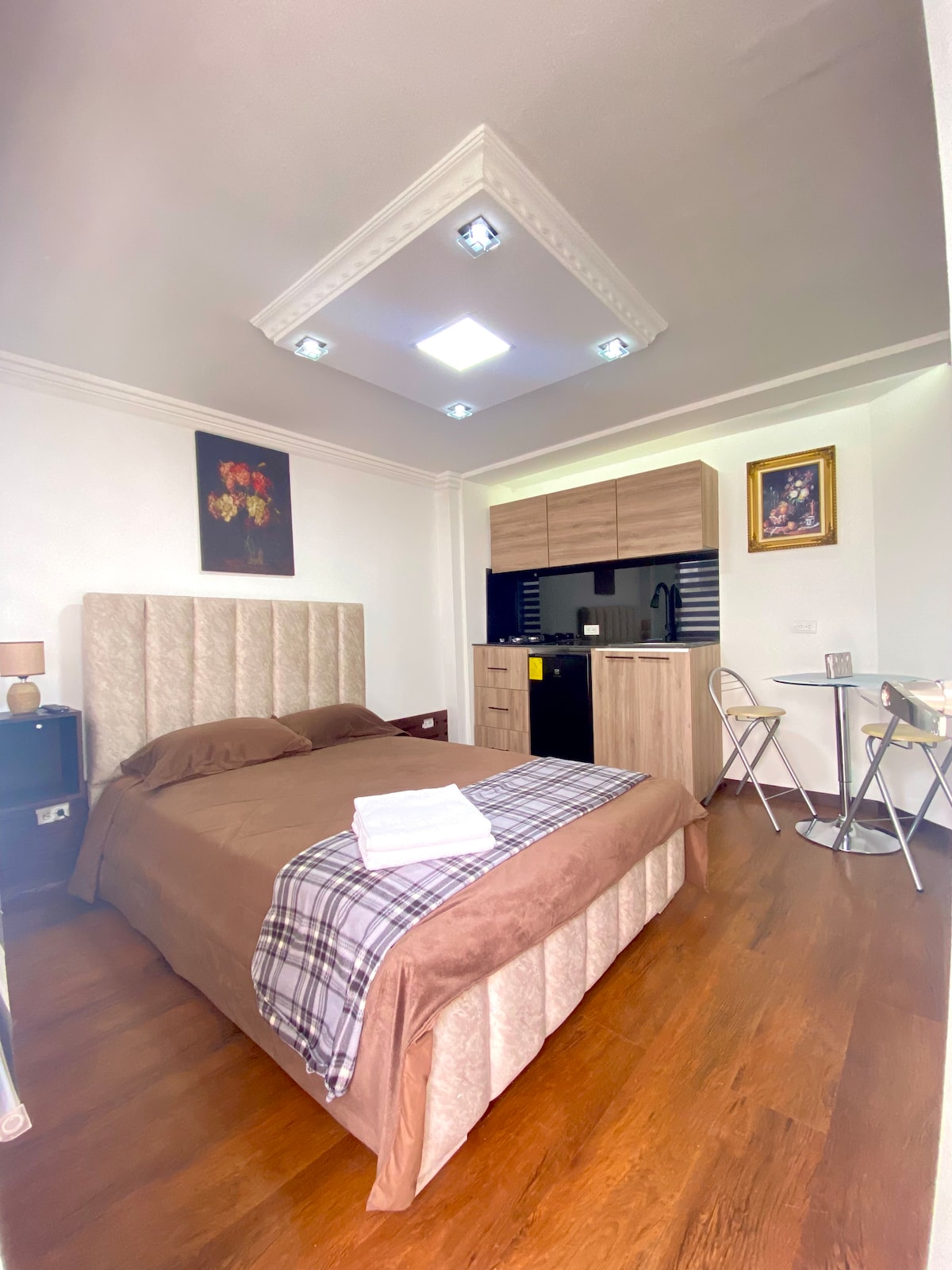 A cozy suite features a spacious bed with a light-colored headboard and a neatly arranged blanket. A small dining table and two chairs are positioned beside the kitchenette, which is equipped with wooden cabinetry. Soft lighting comes from recessed fixtures and natural light enters from nearby windows.