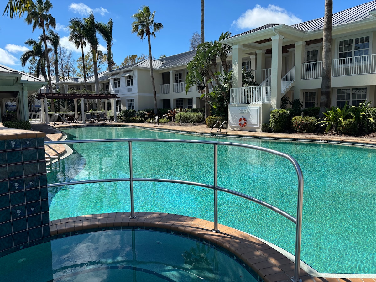 The community pool area features a sparkling blue pool bordered by palm trees and landscaping. A hot tub is visible, positioned next to the pool. The surrounding buildings are painted in soft colors with balconies overlooking the area, creating a relaxing atmosphere.