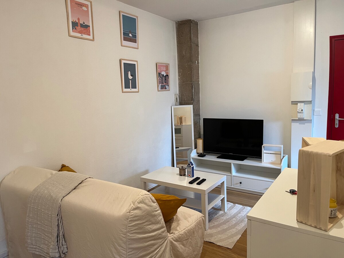 A bright living area features a comfortable sofa with a decorative cushion, facing a flat-screen television. A modern coffee table sits in front, accompanied by a full-length mirror. Light walls are adorned with framed artwork, while the space is completed with a simple rug and an accent table.