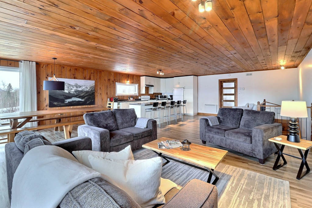 A spacious living area features a combination of comfortable sofas and armchairs arranged around a wooden coffee table. A long dining table can be seen in the background, accompanied by modern bar stools. Soft natural light filters through large windows that enhance the wood-paneled ceiling.