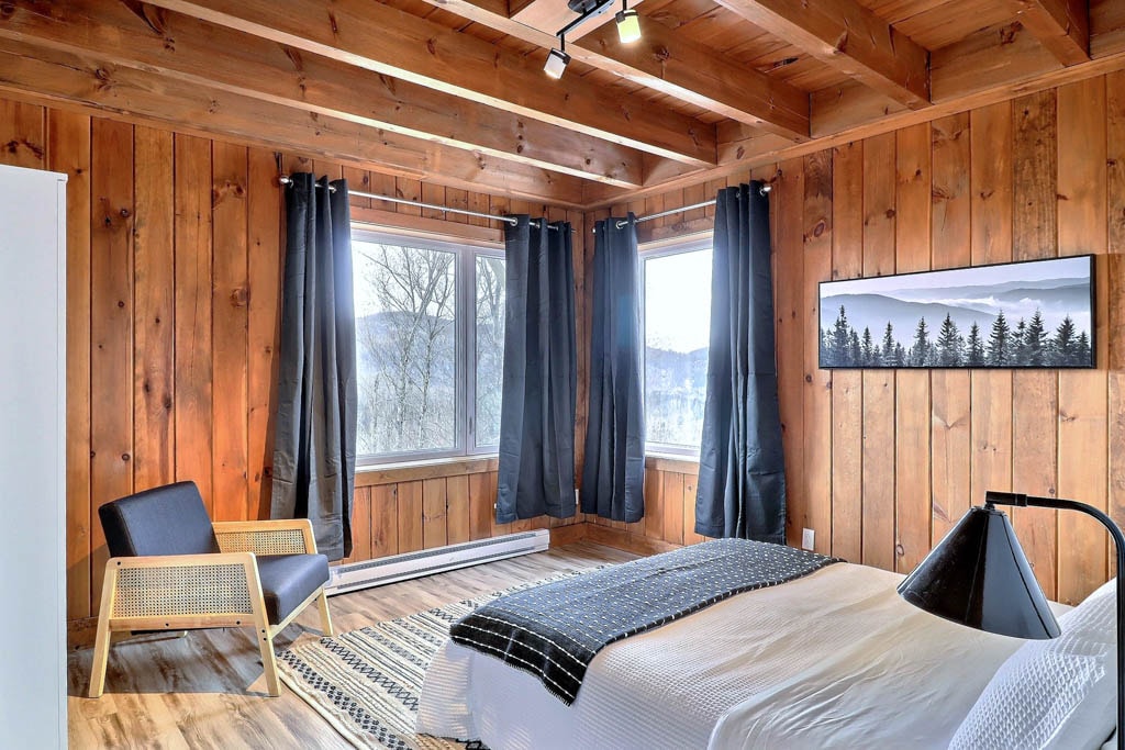 A cozy bedroom is displayed, featuring warm wooden paneling and natural light from two large windows. A light-colored bed with a textured blanket is situated beside a minimalist black lamp. A comfortable chair is positioned nearby, adding to the inviting ambiance of the space.