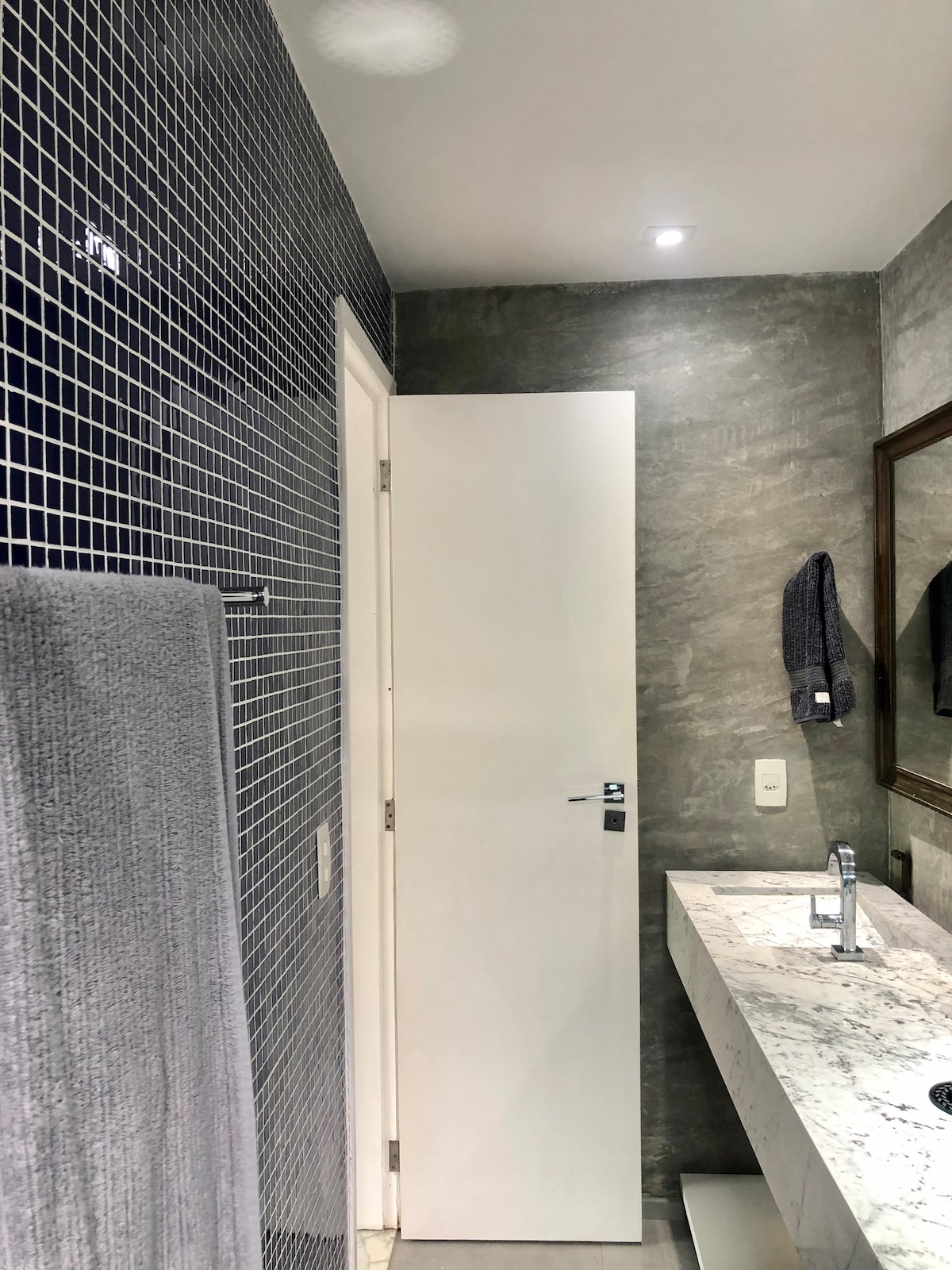A modern bathroom features a sleek, marble countertop with a chrome faucet positioned above a white base. The wall is covered in dark blue tiles, enhancing the contemporary design. A towel hangs on a rack, and a full-length mirror rests above the countertop. A white door leads to another space.