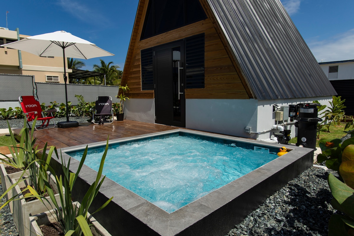 A charming hot tub is located outside a wooden chalet, surrounded by lush greenery. Sun loungers sit nearby under a large umbrella. The exterior features a mixture of wooden and metallic elements, with a sleek door adding to the modern aesthetic.