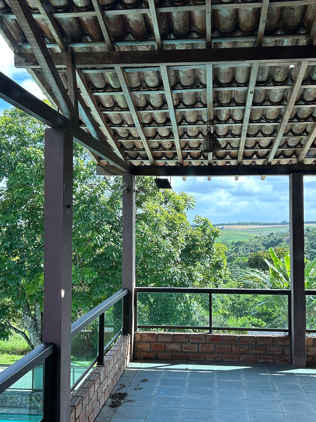 An outdoor covered patio is revealed, featuring a sturdy tiled roof supported by wooden beams. A railing made of glass offers views of lush greenery and distant hills, allowing natural light to fill the space. The textured stone floor adds an element of rustic charm.