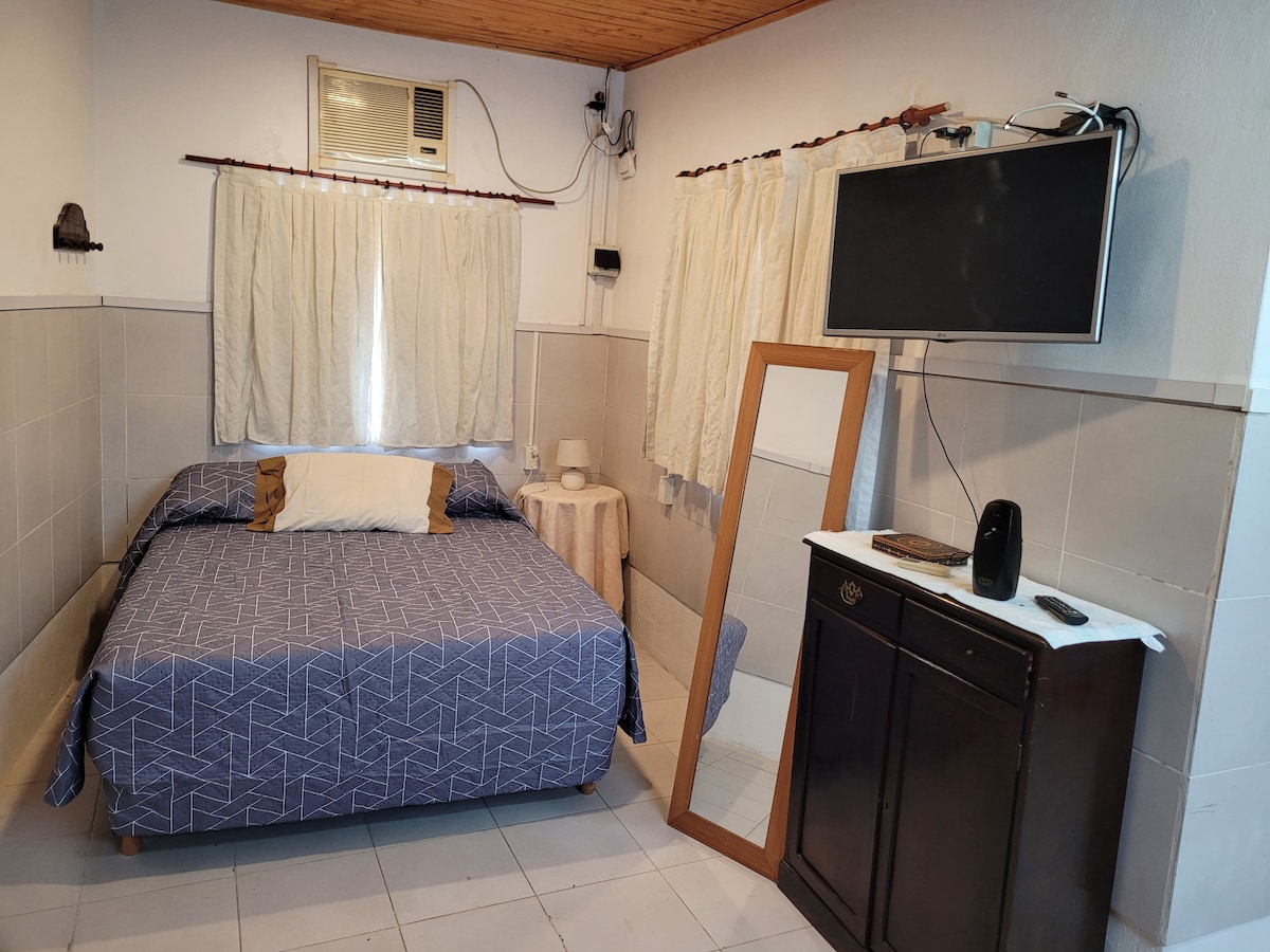 A cozy sleeping area is furnished with a spacious bed covered in a blue geometric patterned blanket. A full-length mirror stands beside a small table, while a television is mounted on the wall. Natural light filters through the white curtains, creating a bright atmosphere.