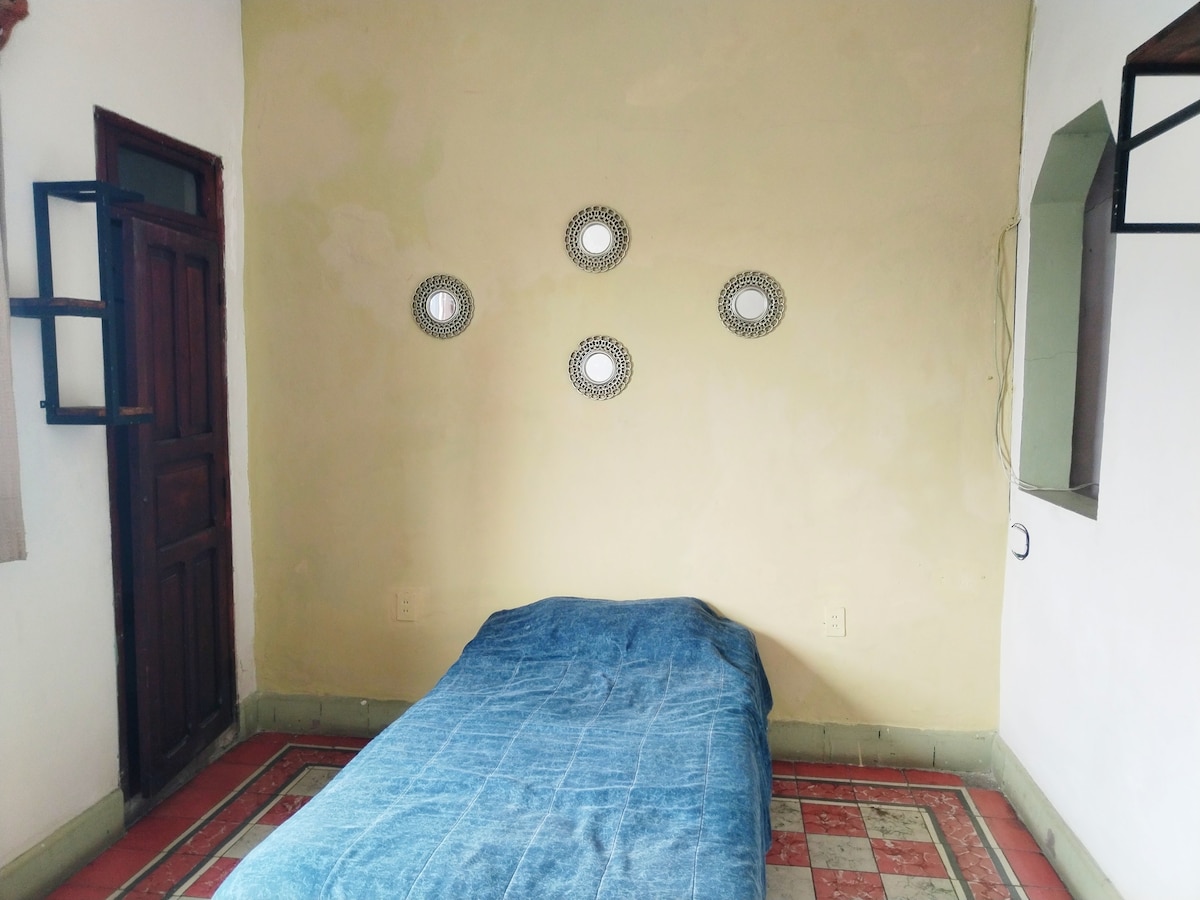 A simple bedroom features a single bed covered with a blue bedspread. Five round mirrors are mounted on the cream-colored wall above the bed. The terracotta tile floor adds warmth to the space, complemented by a wooden door on the left and a window nook on the right.