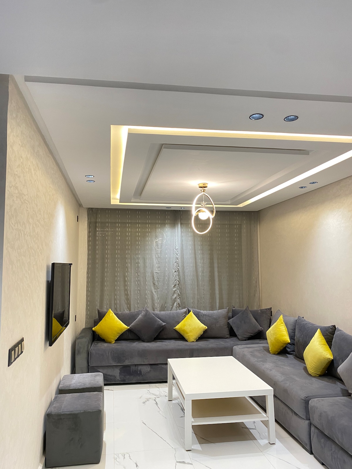 A contemporary living area features a gray sectional sofa adorned with bright yellow cushions. A minimalist coffee table rests in front, with a wall-mounted television providing entertainment options. Soft lighting enhances the room's modern design, while sheer curtains allow natural light to filter in.