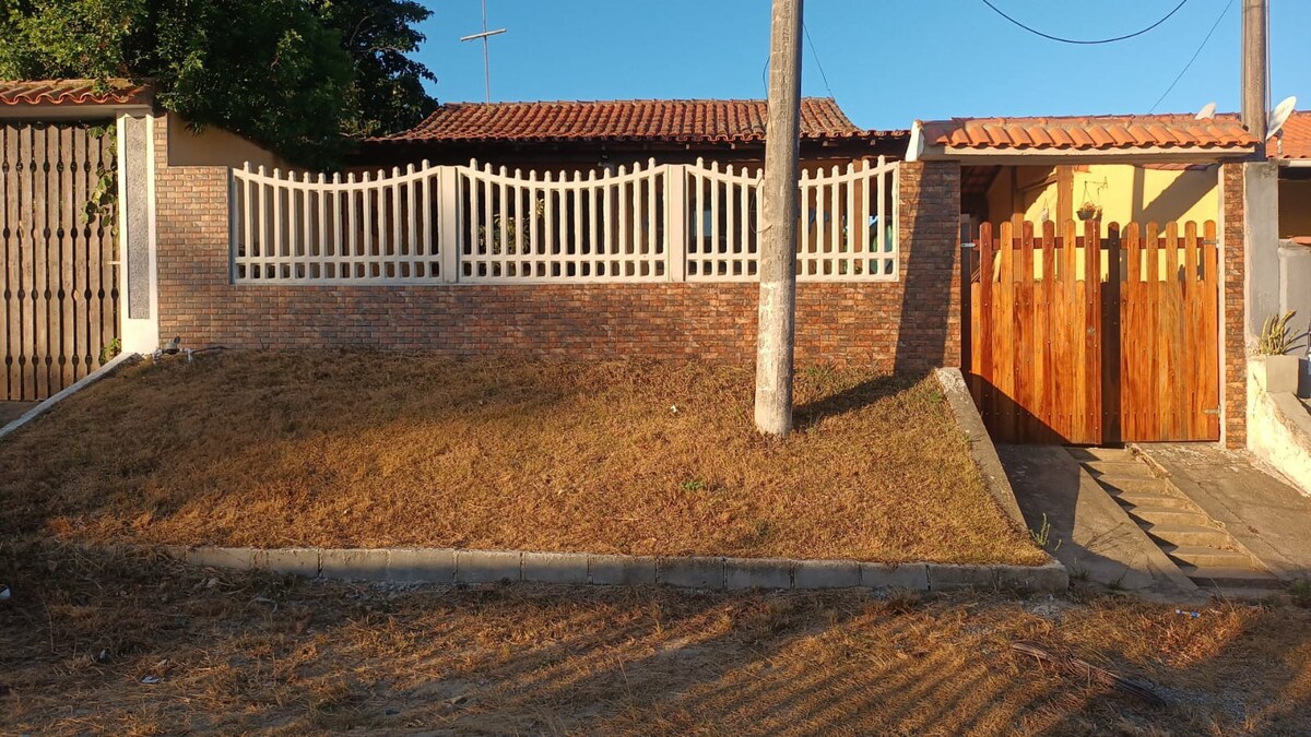 A charming exterior view of a colonial house is presented, featuring a wooden gate and a brick wall. A grassy area slopes gently towards the structure, framed by a tall tree. The terracotta roof contrasts with the clear sky, creating a welcoming entrance.