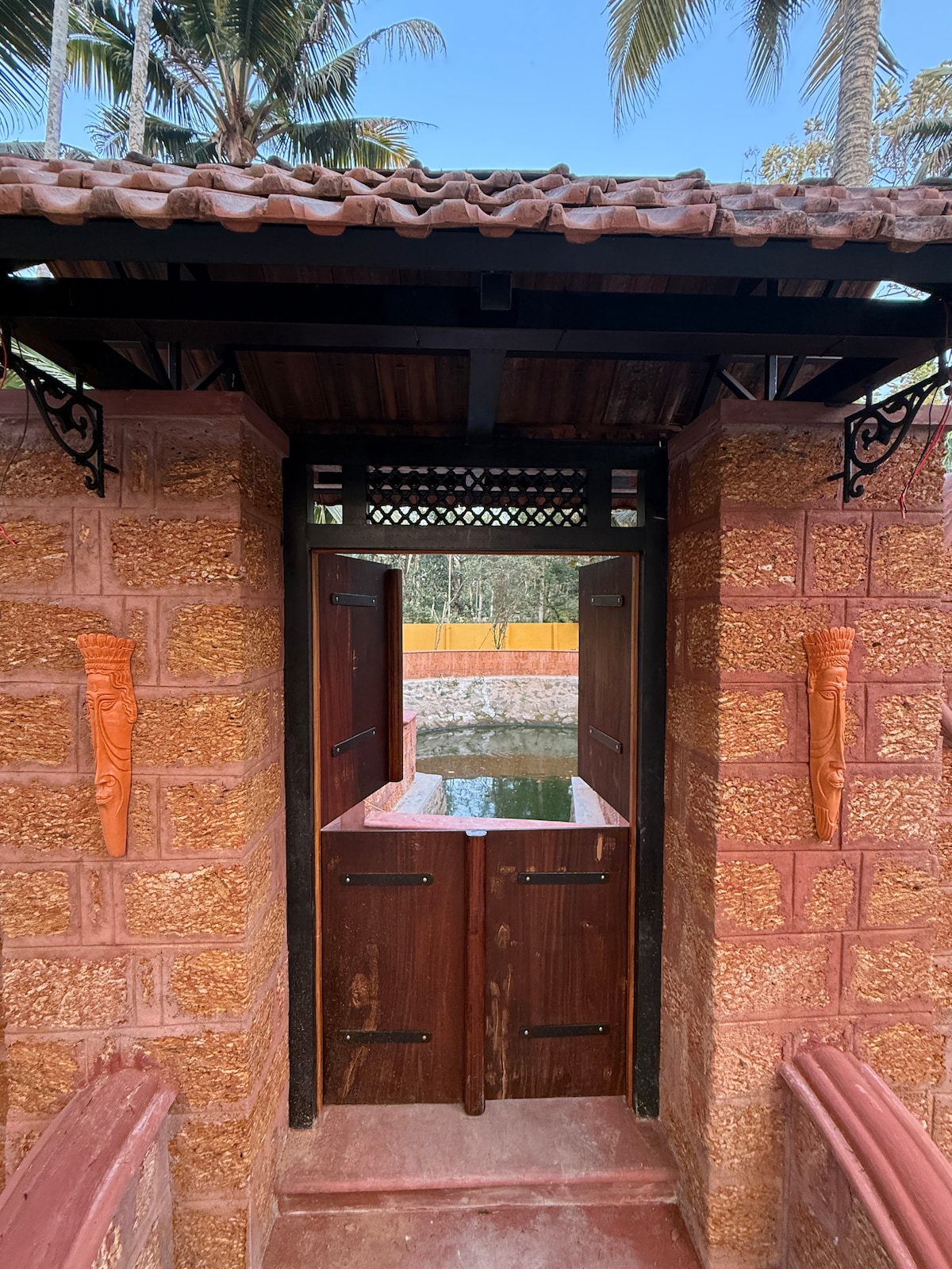 A rustic entrance features two wooden doors, set within textured stone walls. Tropical foliage is visible in the background, framing the peaceful view of a private pond nestled just beyond the entryway.