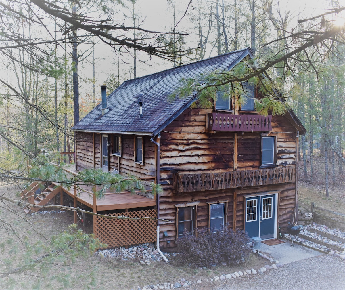 A charming wooden cabin is nestled among tall trees, featuring a spacious deck with seating areas. The exterior showcases natural wooden beams and a balcony, while various windows are visible, allowing light to enter. The area is surrounded by a landscaped yard with gravel pathways.