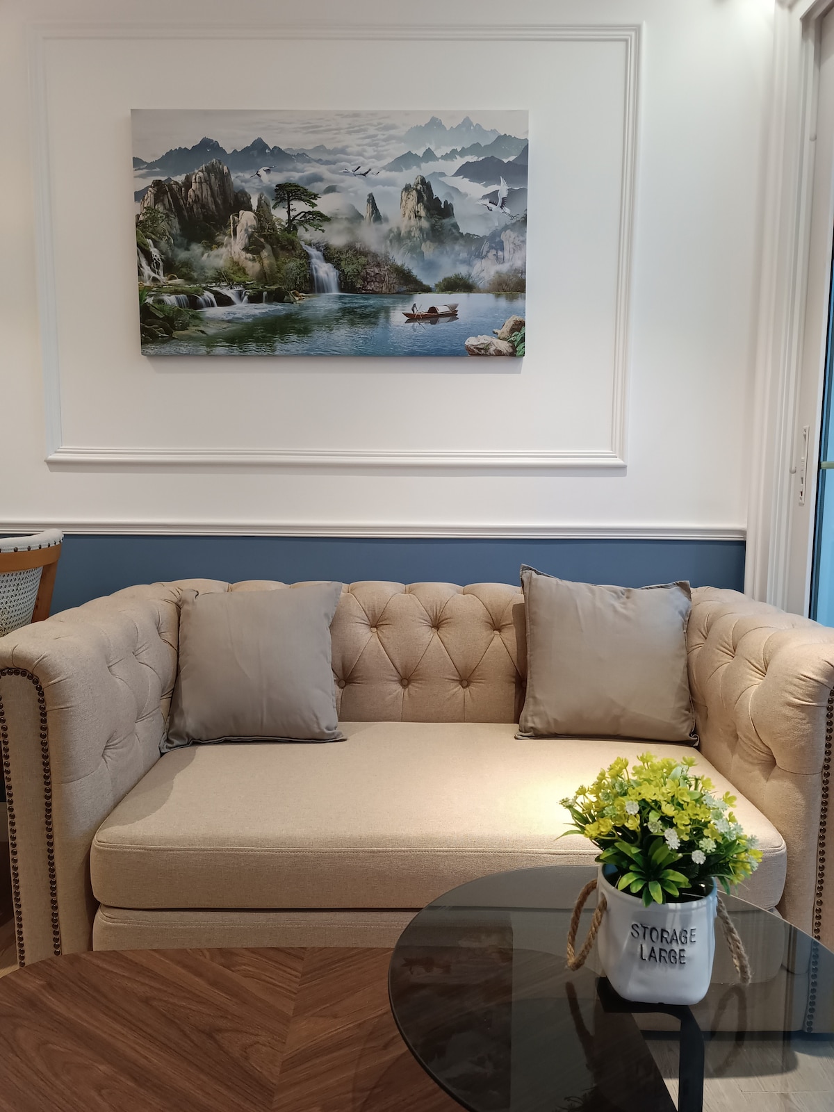 A subtle and elegant living area is depicted, featuring a tufted beige sofa with two soft cushions. Above the sofa, a scenic landscape painting presents mountains and water. A small round table with a decorative plant arranged in a storage container completes the setting, enhancing a cozy atmosphere.