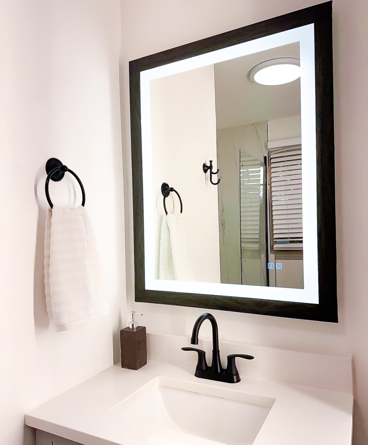 A modern bathroom is depicted, featuring a large mirror framed in black that reflects natural light. A simple sink with a dark faucet is visible, accompanied by a beige towel hanging on a ring. A soap dispenser and a sleek hook add to the minimalist design.