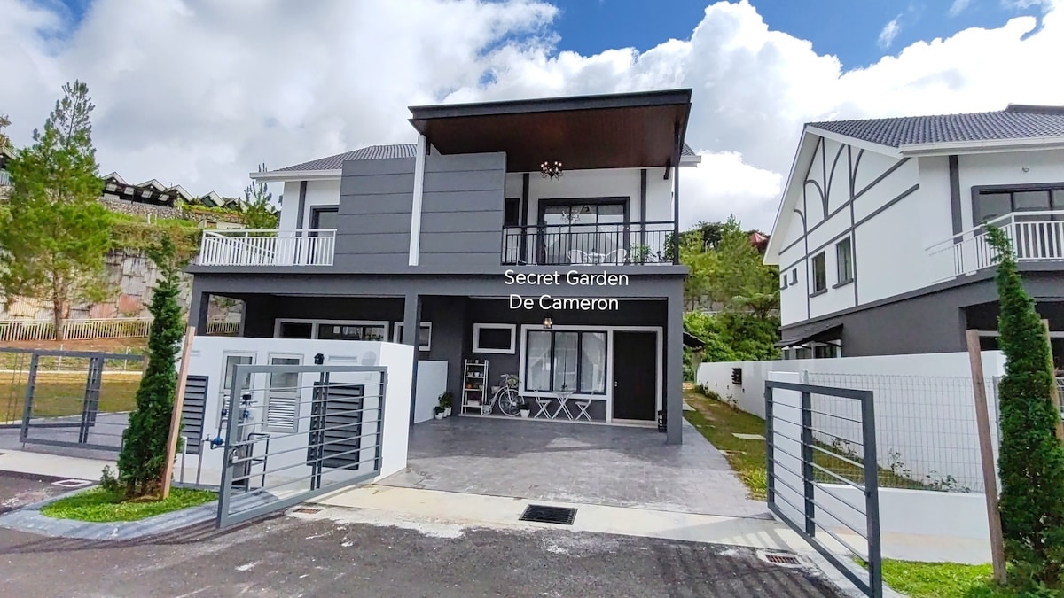 A modern two-story villa, featuring a sleek exterior with horizontal grey and white panels. Large glass doors provide visibility into the welcoming entryway, which offers two chairs and a small table. The surrounding landscape includes greenery, enhancing the fresh atmosphere.