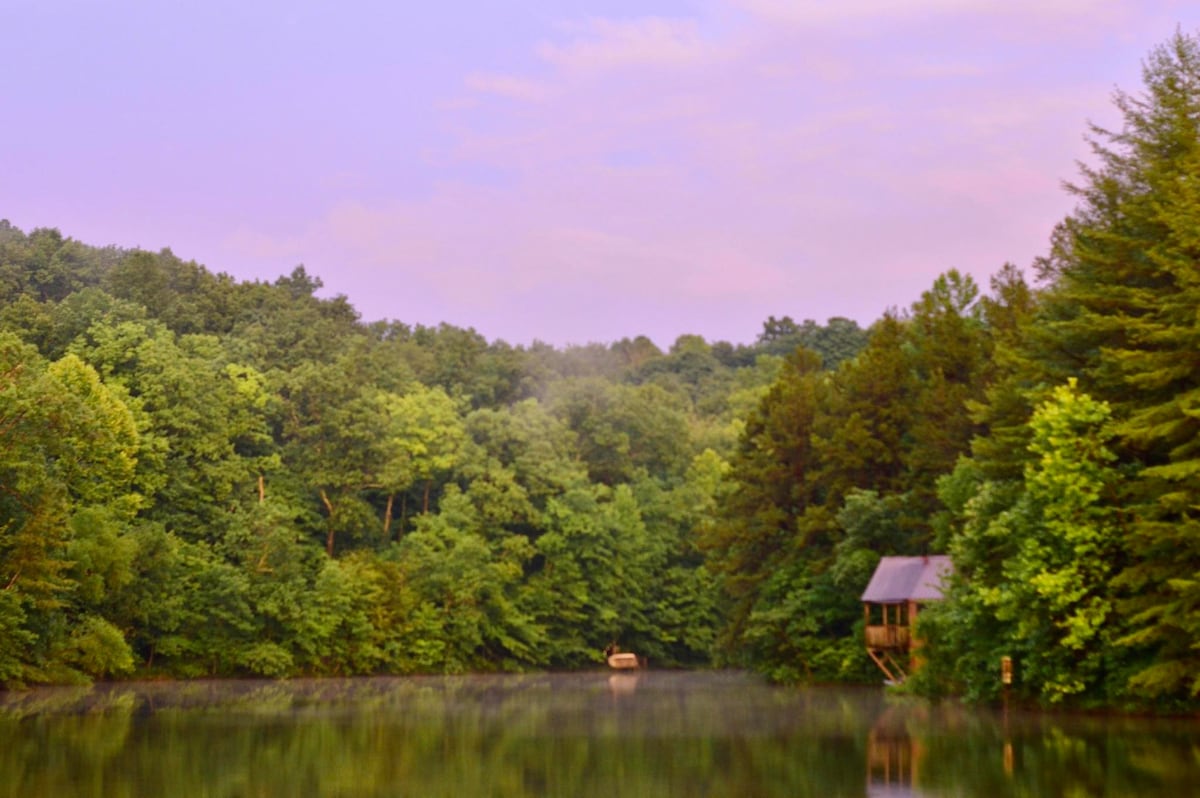 Surrounded by lush greenery, the calm lake reflects the soft colors of dawn. A small boat is moored at a dock, with a rustic cabin visible among the trees on the right. The serene atmosphere invites relaxation and appreciation of nature.