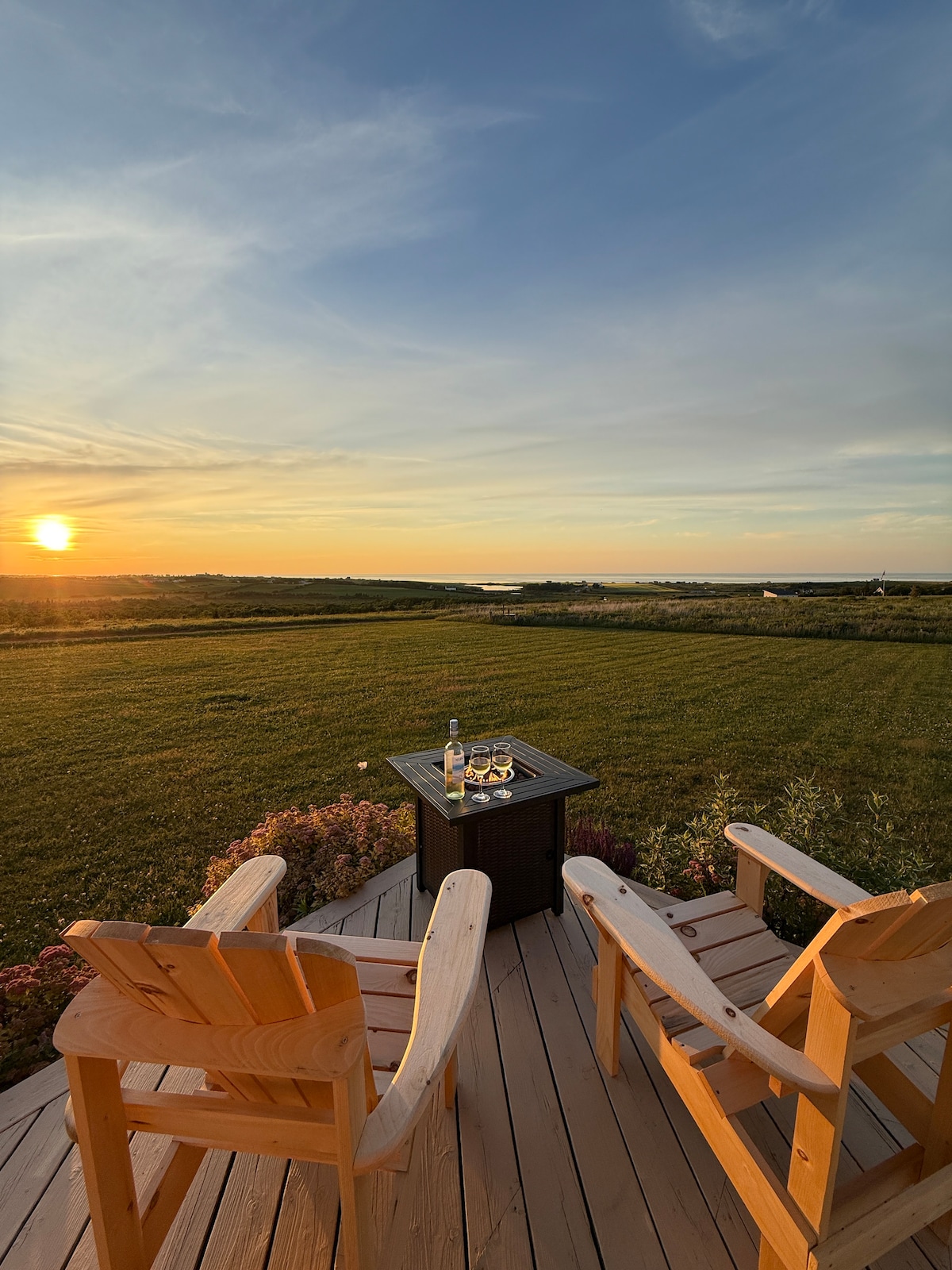 Two wooden adirondack chairs are positioned on a deck, facing a vibrant sunset over a wide open landscape. A small table between the chairs holds a bottle and glasses, with lush greenery visible in the background.