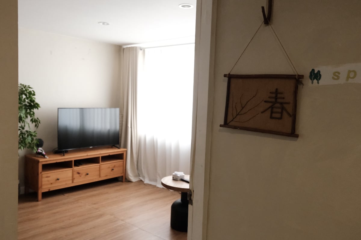 A bright living area is visible through an open doorway, featuring a flat-screen TV on a wooden media console. Natural light filters in through sheer curtains, illuminating a potted plant and a circular table nearby. The warm wood flooring enhances the inviting ambiance.