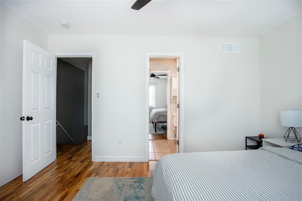 A well-lit bedroom features a spacious bed with a striped duvet. A door leads to a bathroom area, while another door opens to a hallway. Light wood flooring adds warmth to the space, complemented by a small bedside table with a lamp.