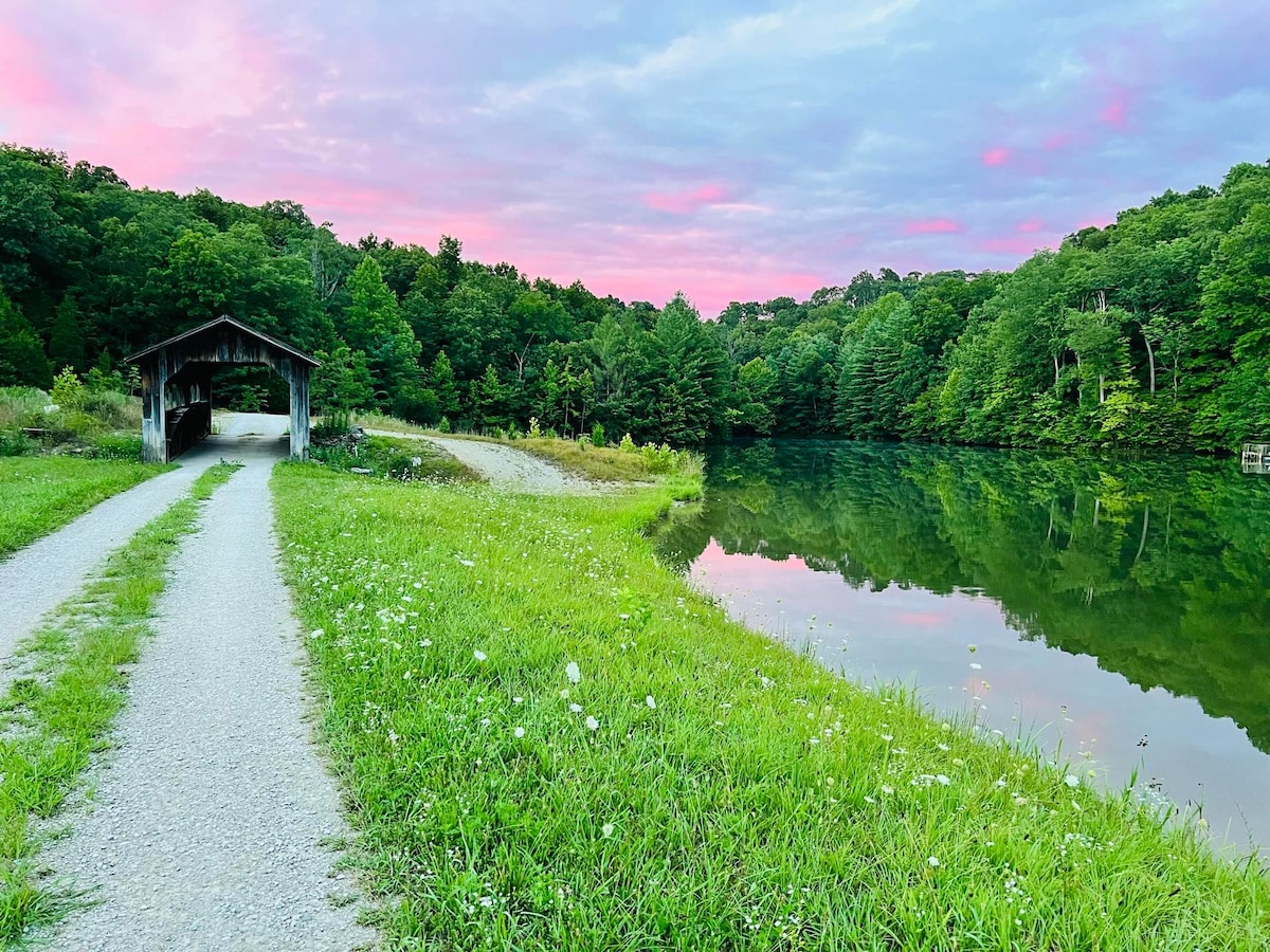 A serene lakeside view is presented, featuring a gravel path leading toward a covered bridge. The calm water reflects the vibrant hues of the sky at dusk, surrounded by lush greenery and scattered wildflowers along the shoreline.