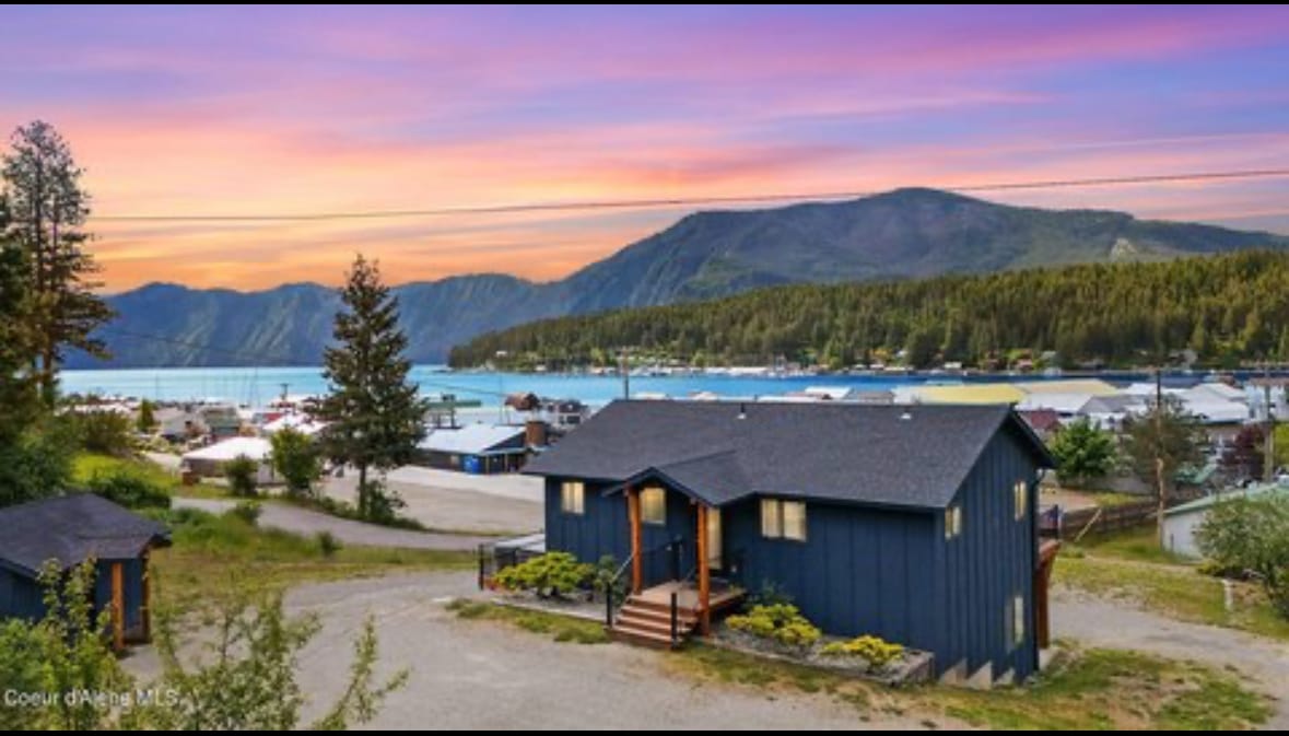 A serene lakeside home is depicted against a colorful sunset sky. The structure exhibits a modern design, with dark exterior siding and multiple entrance steps. Surrounding trees and views of Lake Pend Oreille complement the tranquil setting, while the distant mountains add depth to the landscape.