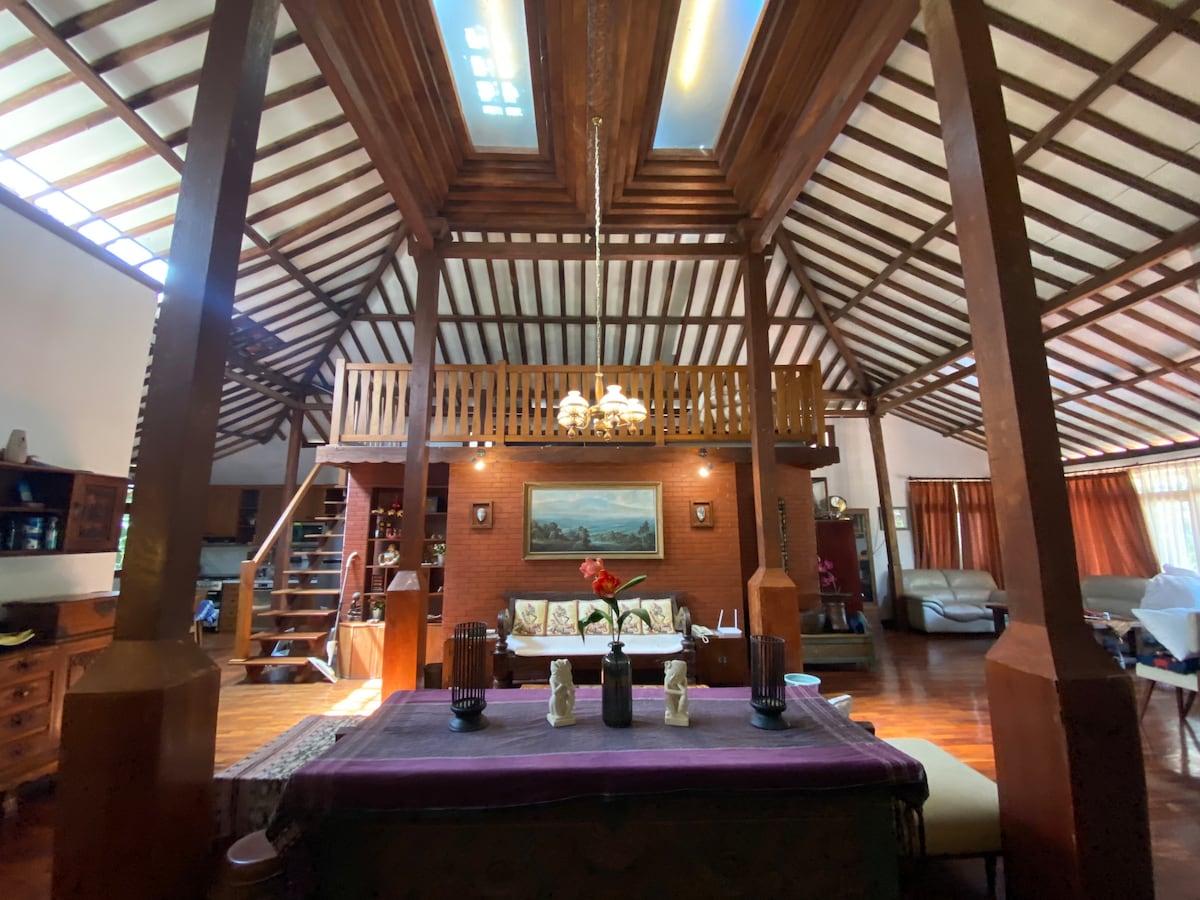 The interior space features high wooden ceilings with skylights, allowing natural light to filter through. A cozy seating area is visible, with a staircase leading to a lofted level. Decorative elements include floral accents and artworks adorning the walls.
