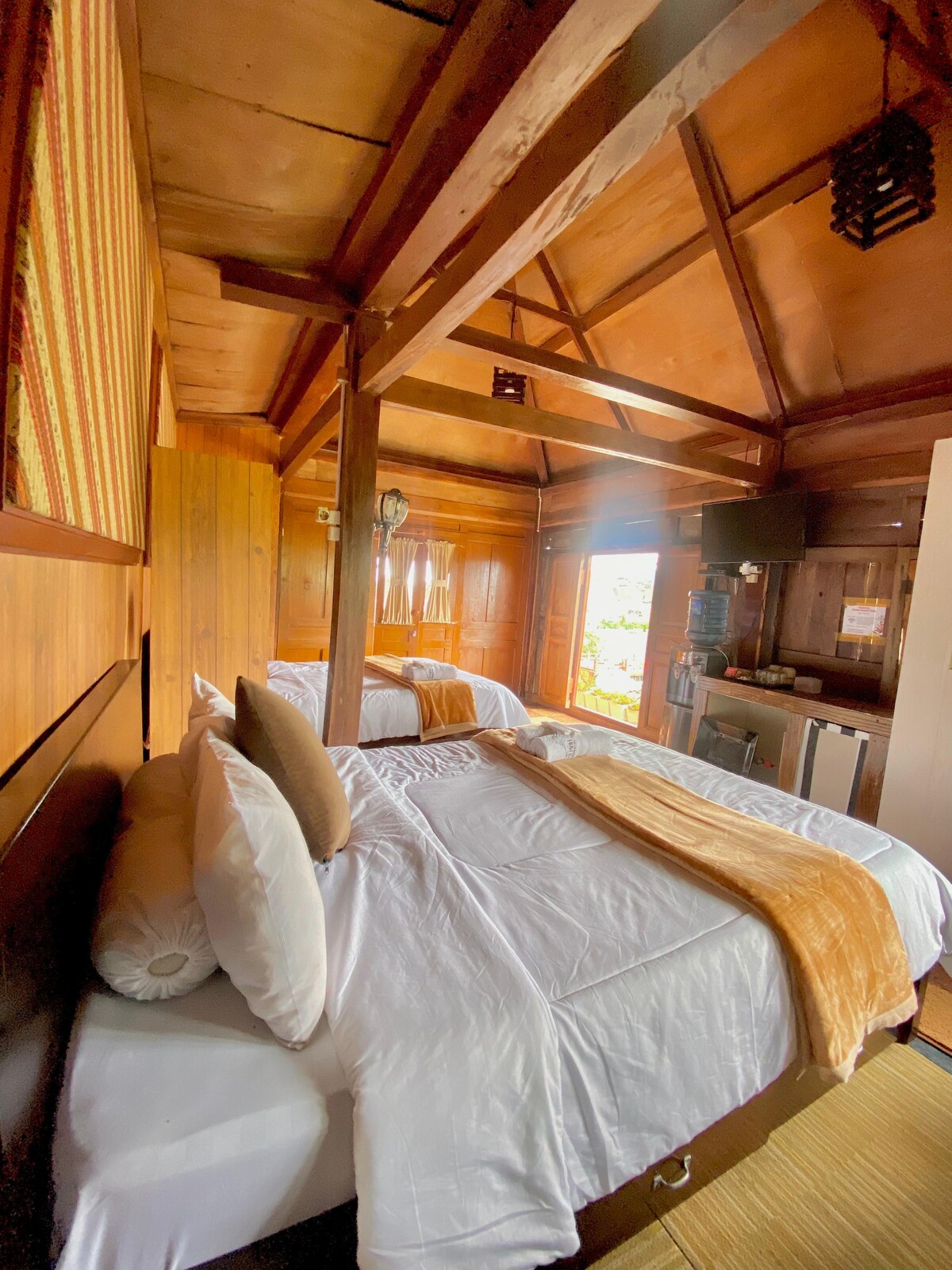 A wooden room features two spacious beds draped in white linens, with an orange throw blanket on each. Natural light is welcomed through large windows, while a television and small refrigerator are positioned against the wall. The ceiling showcases exposed wooden beams.
