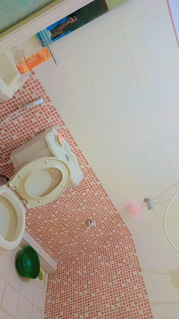 The bathroom features a tiled wall with a mosaic pattern in warm tones. A toilet and a shower area are present, with toiletries arranged visibly. A green basin sits on the floor, completing the functional design of the space.