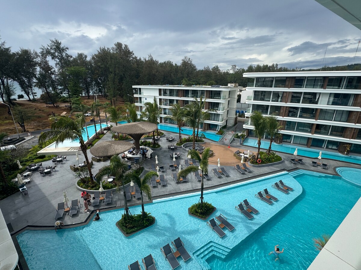 A panoramic view of the resort’s pool area features multiple swimming pools, surrounded by lounge chairs and palm trees. The serene layout creates inviting spaces for relaxation, while various pathways lead to the nearby buildings. The beach and natural surroundings are visible in the background.