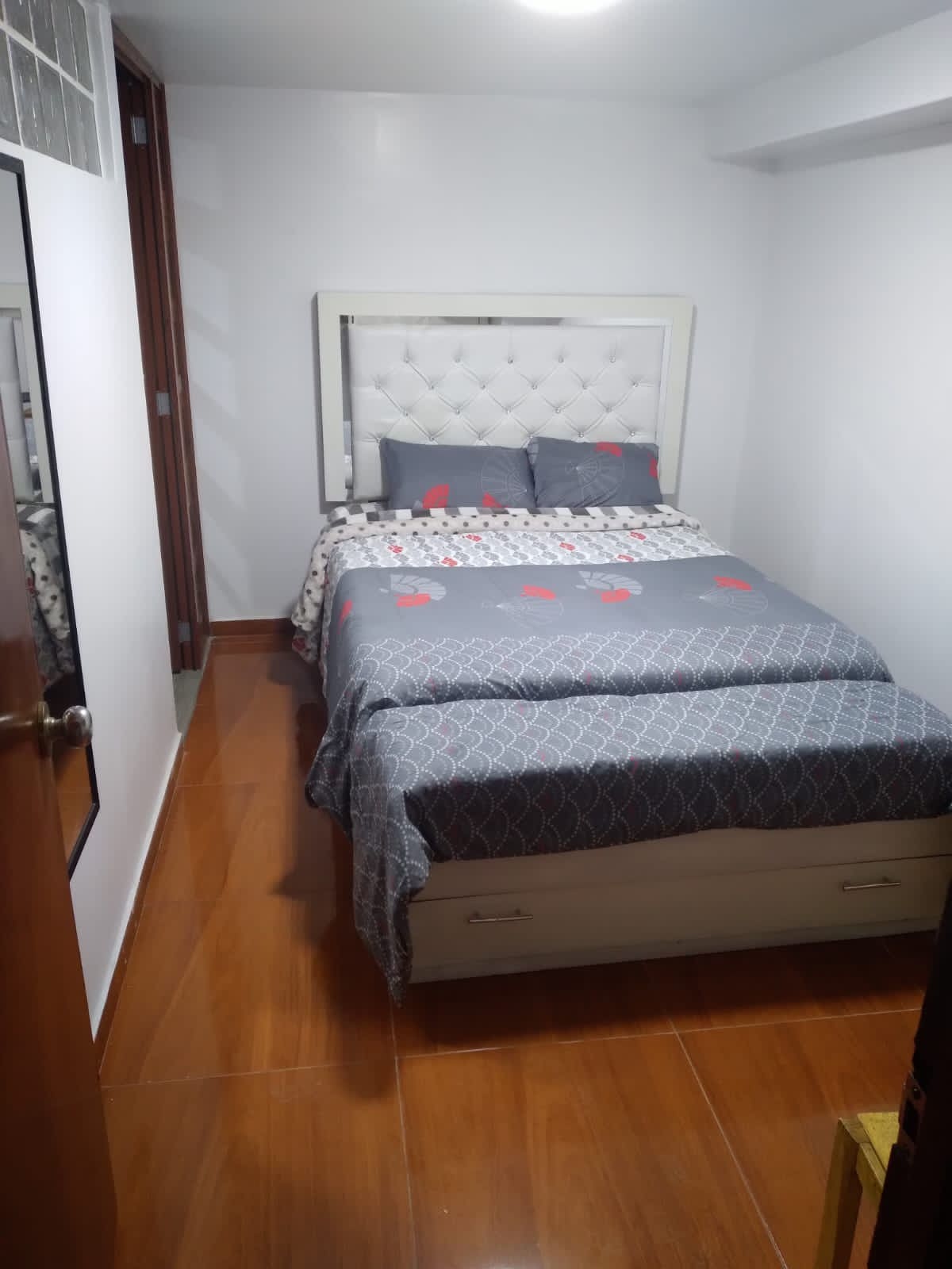 A neatly arranged sleeping area is displayed, featuring a compact bed with a light grey and red patterned cover. Soft pillows rest against the headboard. The floor is tiled in a warm tone, and a mirror reflects the room's layout, enhancing the sense of space.