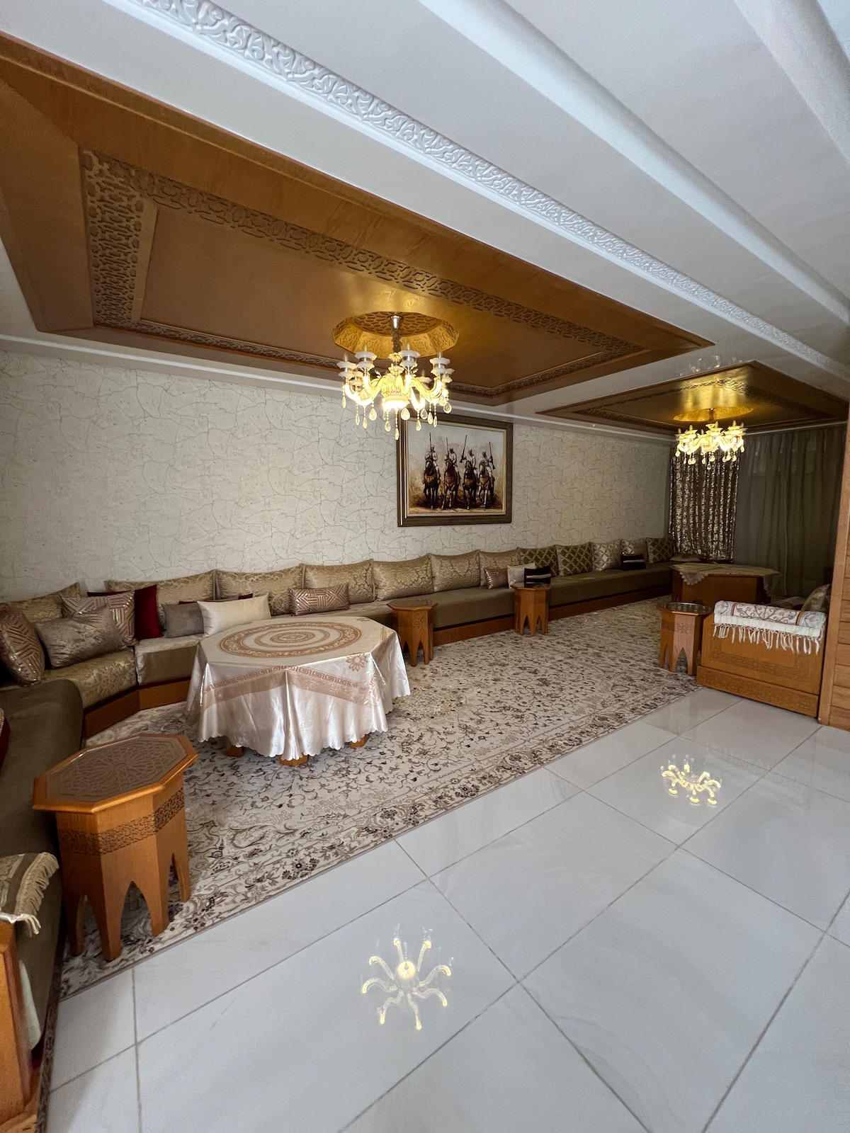 A spacious living area is furnished with elegant seating arrangements and a round table adorned with a tablecloth. Decorative lighting fixtures hang from the ceiling, complementing the intricate patterns on the walls. A plush carpet covers the tiled floor, adding comfort to the setting.