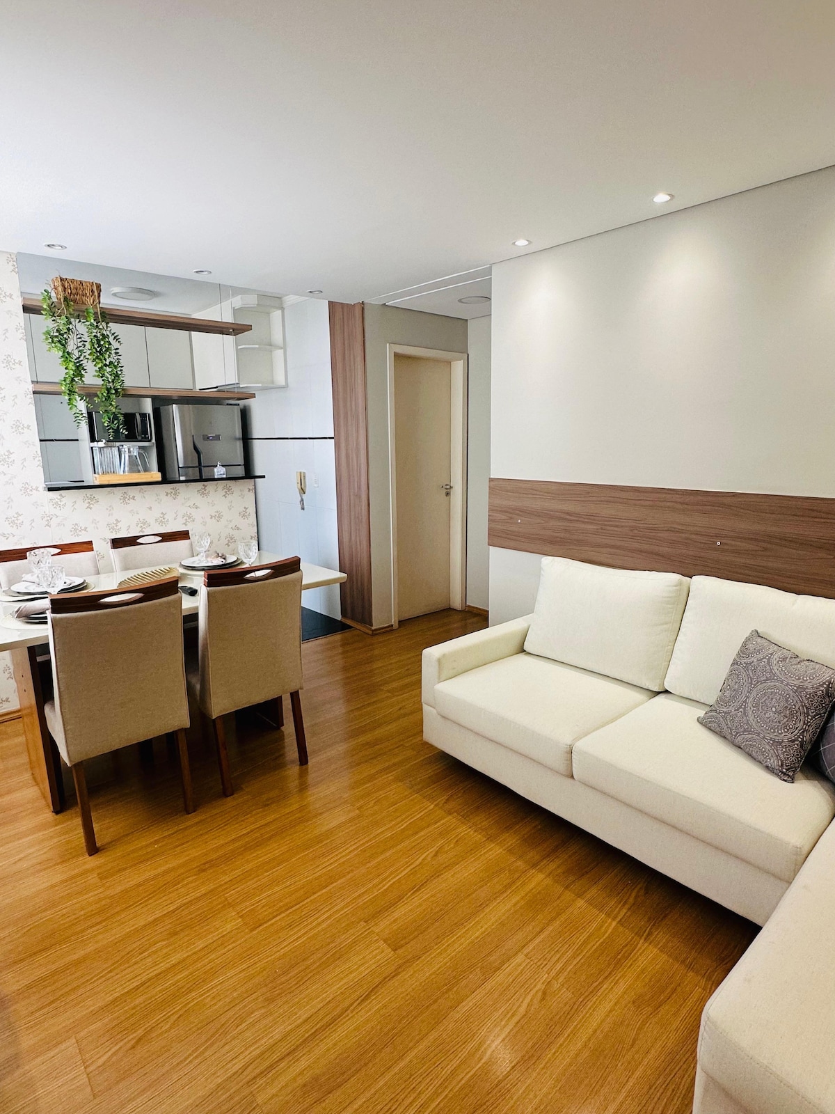 An open living area is highlighted by light wood flooring. A cozy, cream-colored sofa is positioned near a dining table with four chairs. The kitchen features modern appliances, including a refrigerator, and a decorative plant hangs above the counter.