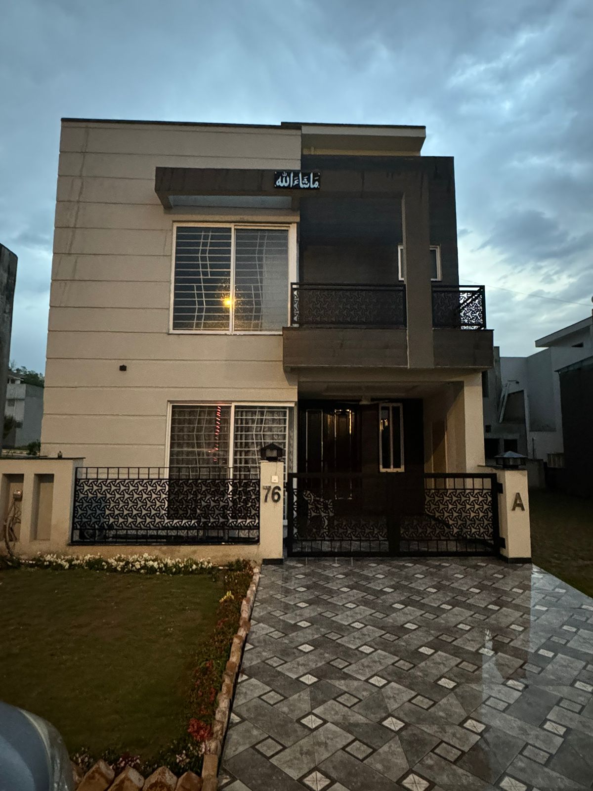 A modern two-story house features a symmetrical façade with large windows and a welcoming entrance. A gated front yard is framed by lush grass and decorative landscaping, while the driveway is composed of patterned tiles, leading to the main entrance.