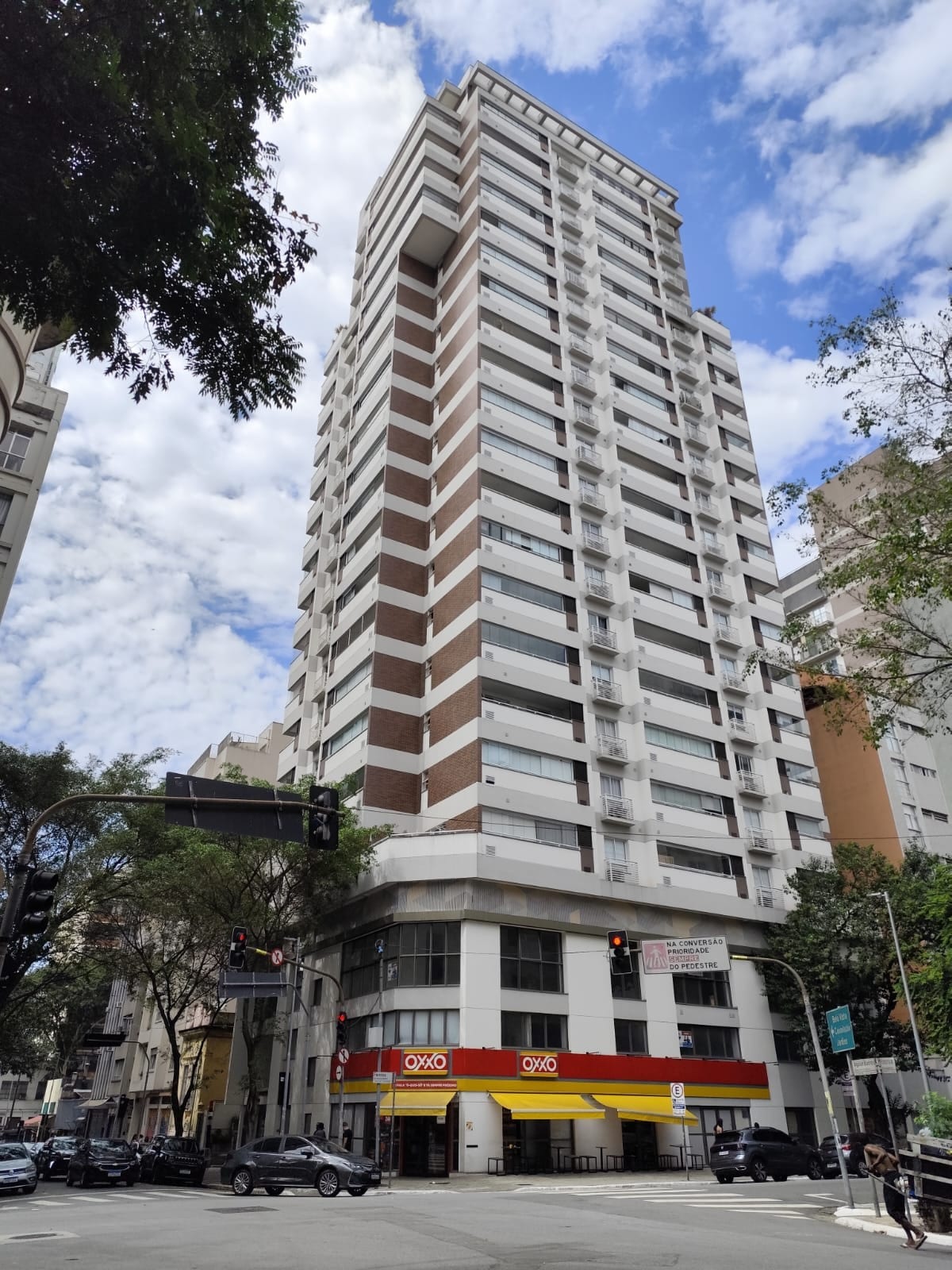 A modern high-rise building is observed from street level, showcasing a mixture of white and brown facade elements. Large windows provide a uniform look across the floors. The street is lined with trees and nearby buildings, contributing to a vibrant urban atmosphere.
