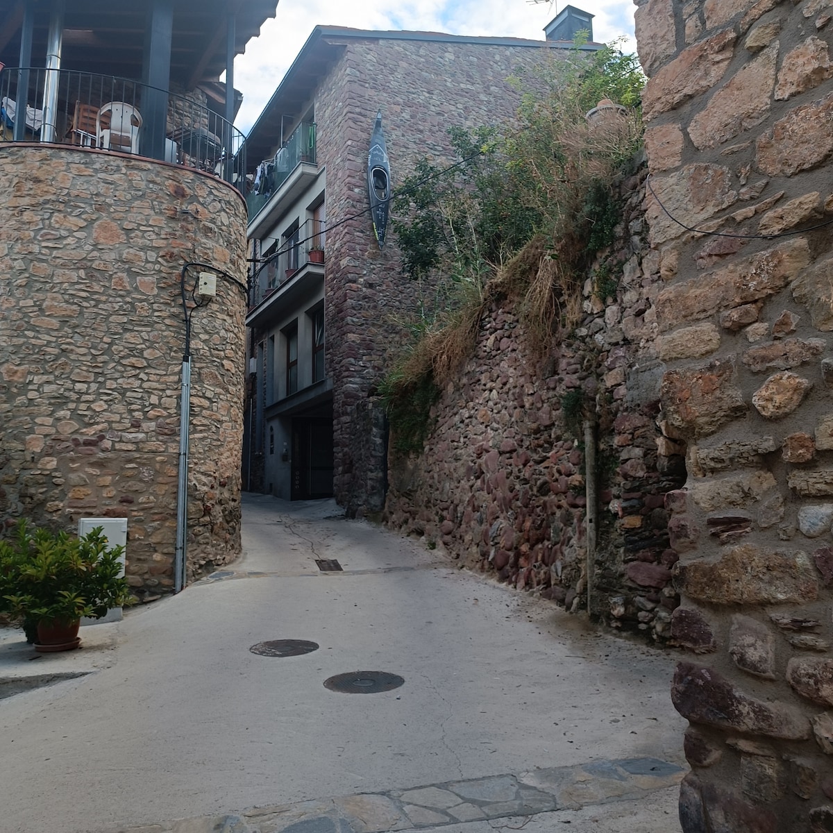 A narrow street is flanked by stone walls, showcasing the rustic architecture of the area. A building with a stone facade is visible in the background, while greenery grows along the walls. The cobblestone path leads into the distance, creating a sense of quaint charm.
