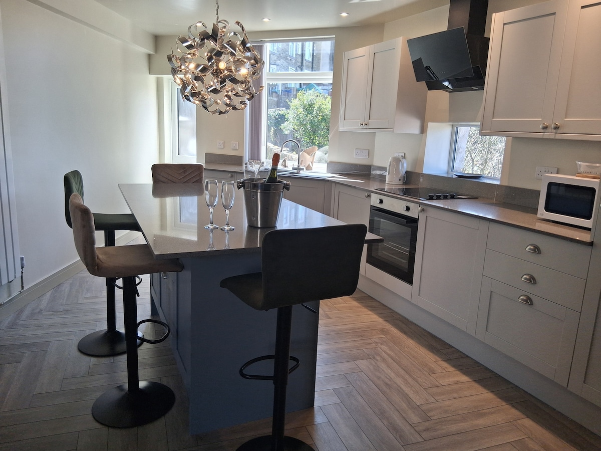 A modern kitchen space is highlighted by sleek cabinetry and a central island with seating. Four bar stools are positioned around the island, while a stylish chandelier adds illumination. Natural light filters through large windows, creating an inviting ambiance.
