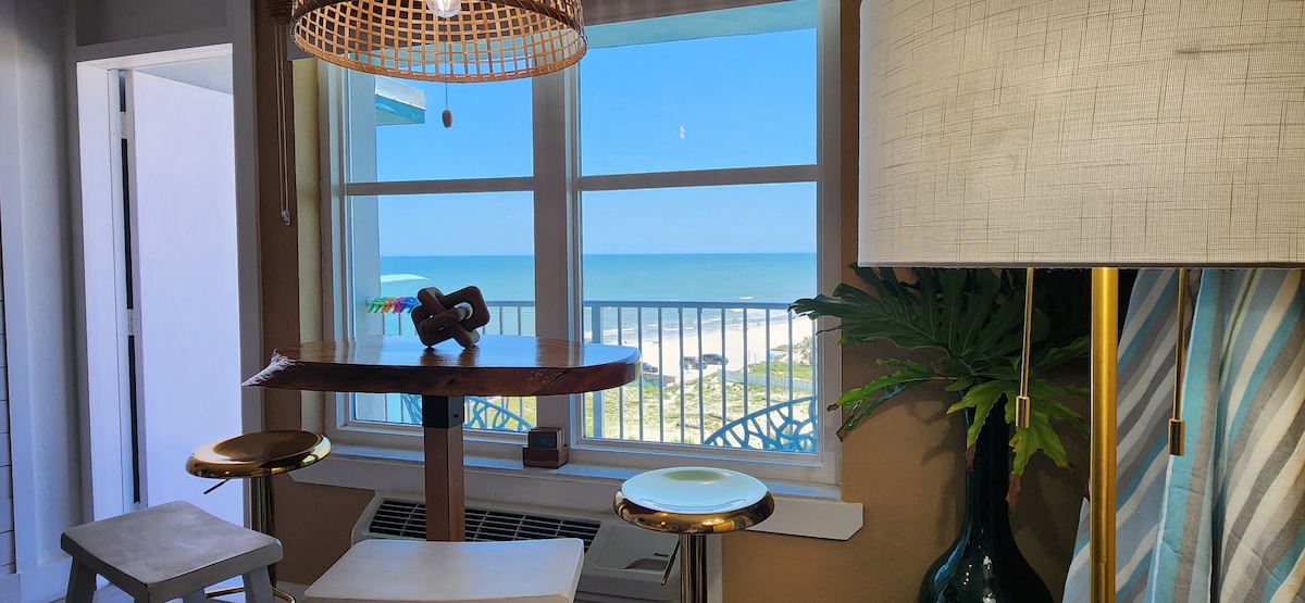 A dining area is positioned beside a large window, providing a view of the ocean and beach. A wooden table with three stools offers seating. A lamp with a textured shade stands nearby, complemented by a potted green plant. The space is filled with natural light.