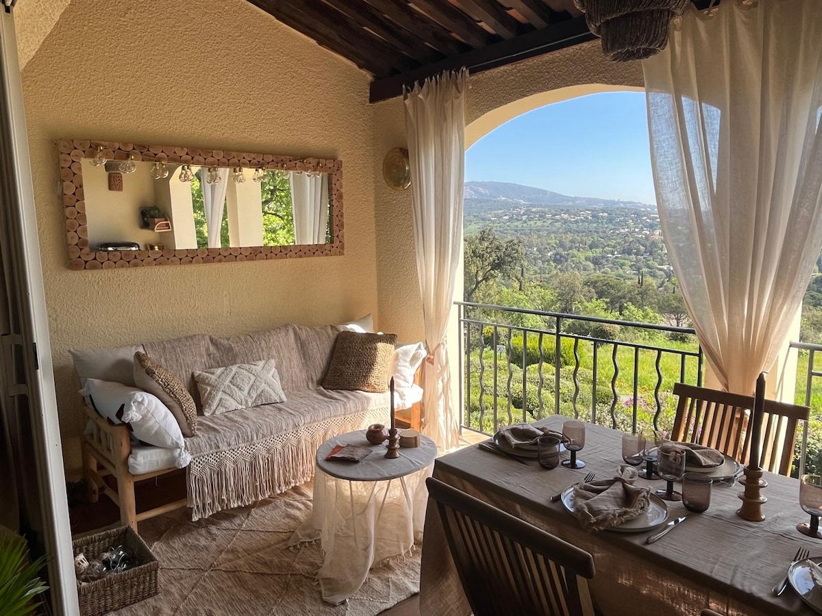 A covered loggia features a comfortable seating area with a textured sofa adorned with plush cushions. A round table is set for dining, surrounded by natural light flowing through sheer curtains, with expansive views of the surrounding hills visible through the archway.