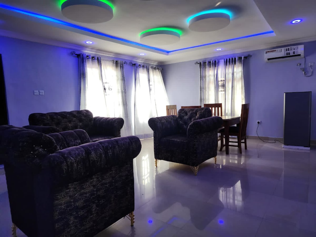 A spacious living area features plush sofas with a dark upholstery, accompanied by a wooden dining table and chairs. Natural light filters through large windows adorned with sheer curtains. The ceiling showcases modern recessed lighting, providing a calming ambiance.