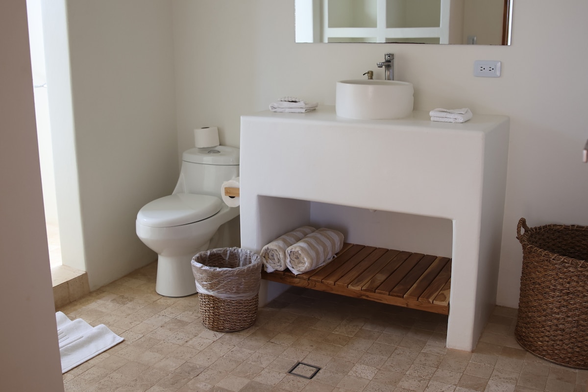 A modern bathroom features a white pedestal sink above a wooden shelf stocked with rolled towels. A toilet is positioned beside the sink. The floor is covered with neutral-toned tiles, and a wicker basket and a waste bin are placed on the floor for additional storage.