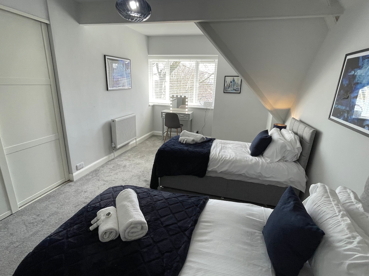 A well-lit bedroom features two adjustable beds, dressed in white and navy linens. A small desk with a chair is positioned near a window, allowing natural light to fill the space. Soft, neutral carpet adds comfort underfoot.