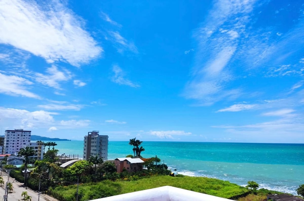 An expansive view of the ocean is captured under a bright blue sky, showcasing the vibrant turquoise water. Palm trees and buildings line the coastline, providing a sense of the surrounding area. The image highlights a peaceful seaside environment.