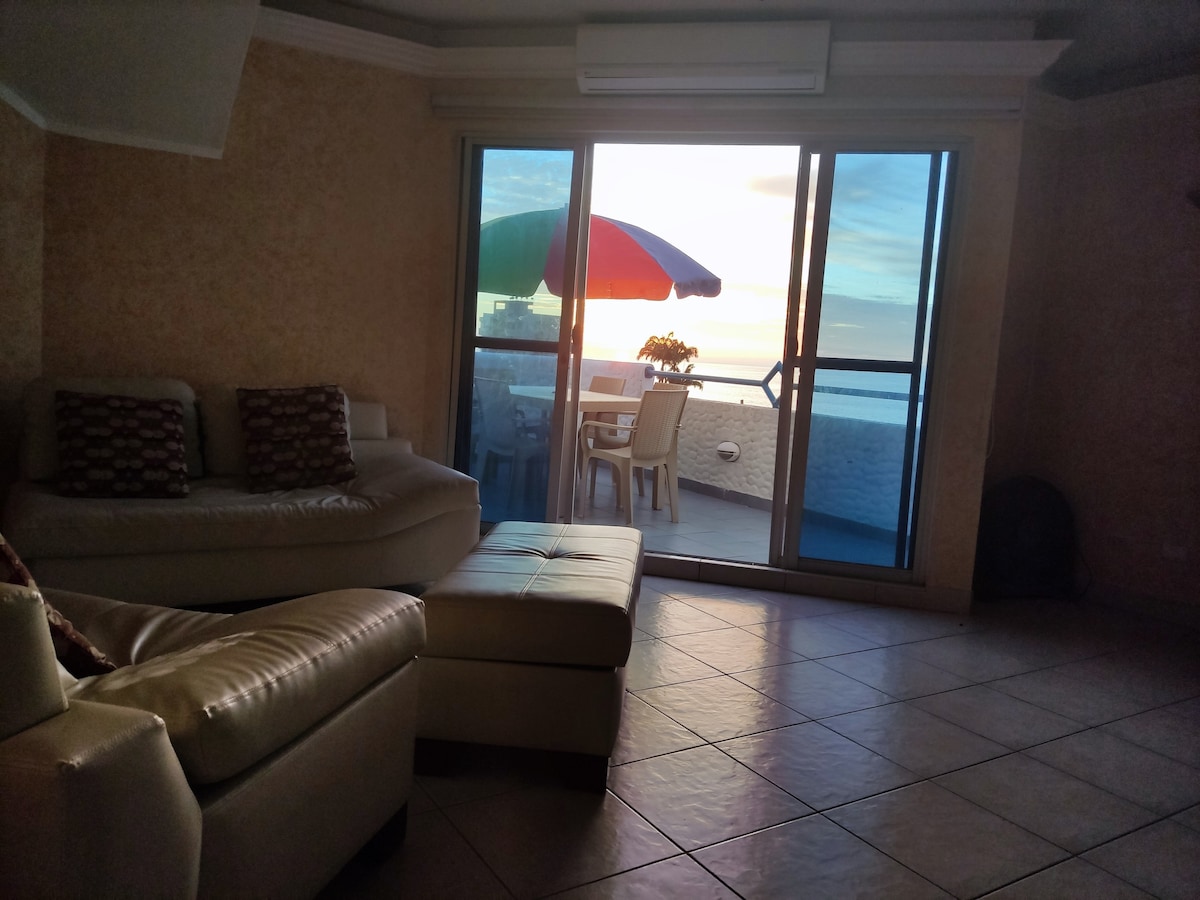 A spacious living area features a large sectional sofa, positioned opposite a glass sliding door. The door opens to a balcony with a table and chairs, offering views of a vibrant sunset and colorful umbrella. Soft natural light fills the room.