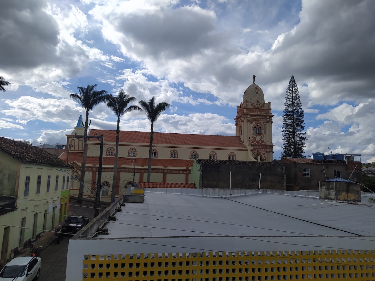 The image captures a view of a historical building with ornate architectural details, complemented by a clock tower. Lush palm trees frame the scene, and a mix of clouds is visible in the sky, suggesting a well-lit outdoor environment enhanced by varied textures.