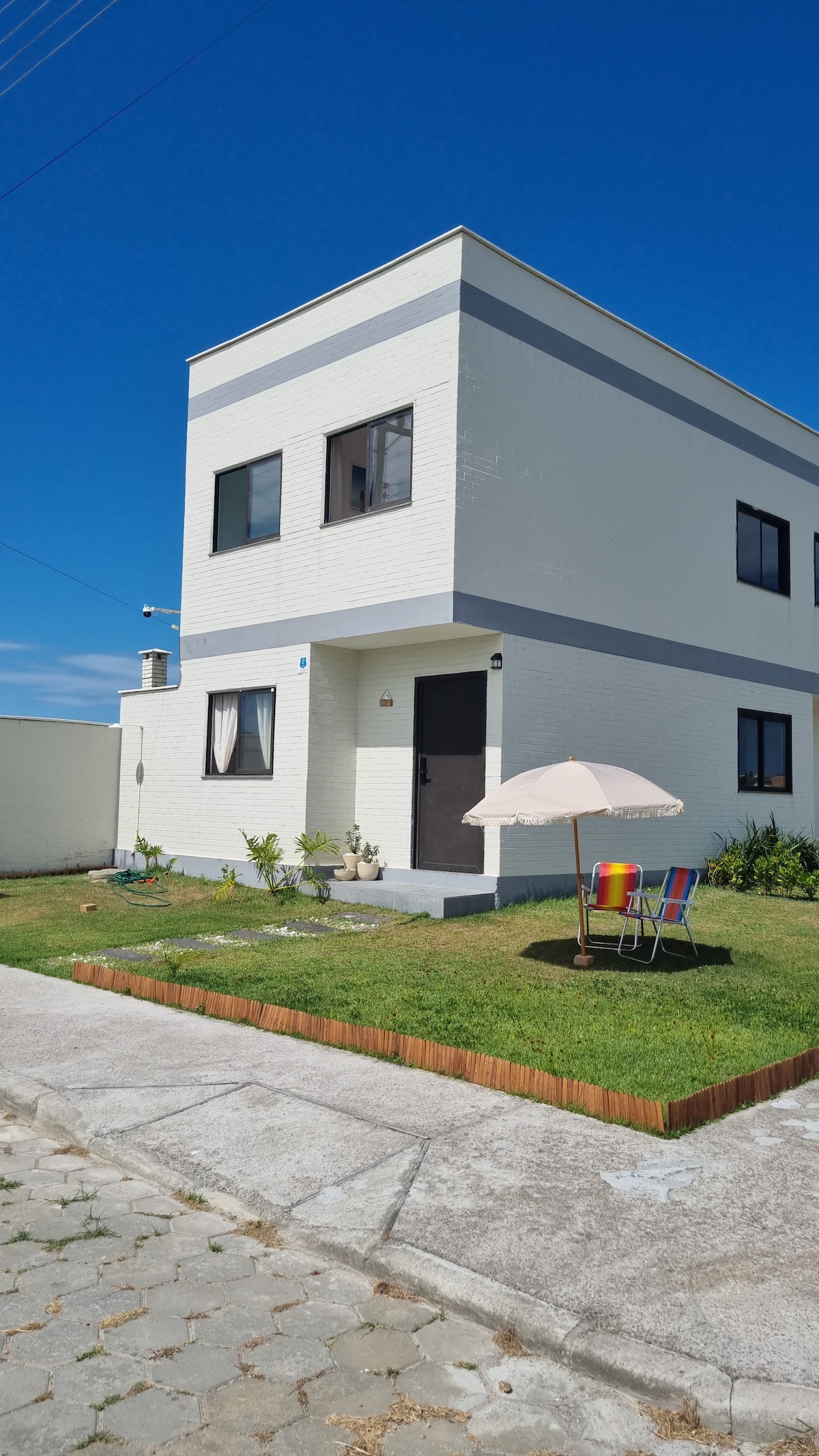 A modern two-story house is presented with a light-colored exterior and large windows. A small seating area with colorful chairs and an umbrella is situated on the well-manicured lawn. Clear blue skies provide a bright backdrop to the inviting front entrance.