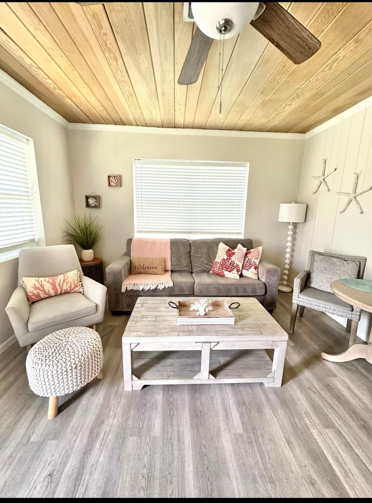A bright living area features a gray sofa adorned with decorative pillows, alongside a light-colored armchair and a textured ottoman. A wooden coffee table is centered in the room, and a side table with a lamp is present. Natural light filters through the window, highlighting the warm wood ceiling.