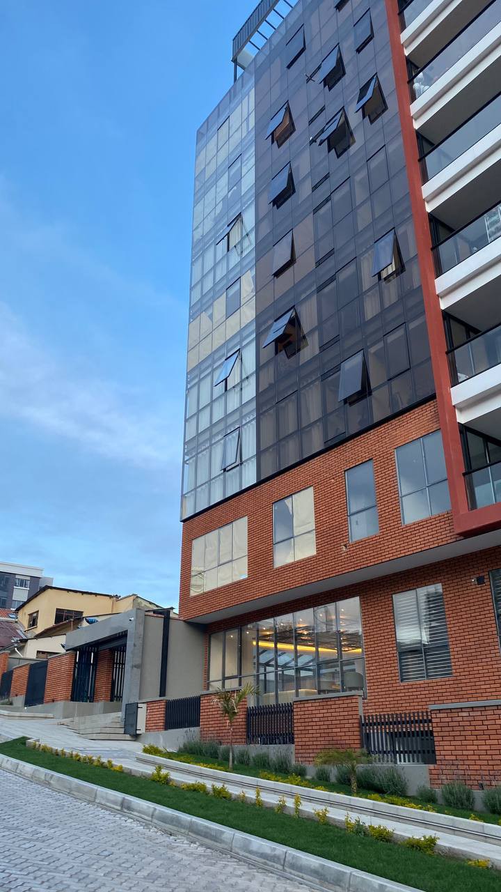 The building stands tall with a modern design, featuring large glass windows reflecting the sky. The lower section showcases a combination of brick and glass, providing an inviting entrance. A pathway lined with greenery leads to the building, highlighting its contemporary architecture.