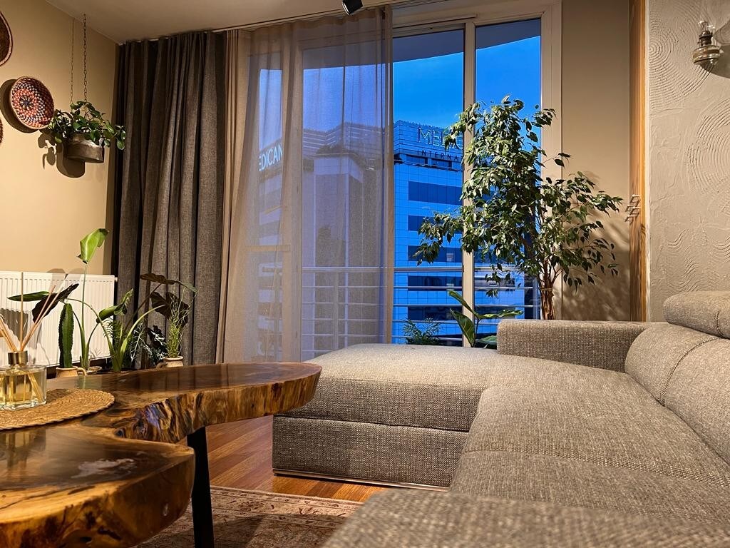 A cozy seating area features a modern, L-shaped gray sofa positioned near a large window with sheer curtains. The view includes nearby buildings. A decorative wooden table and houseplants add a touch of nature to the space.